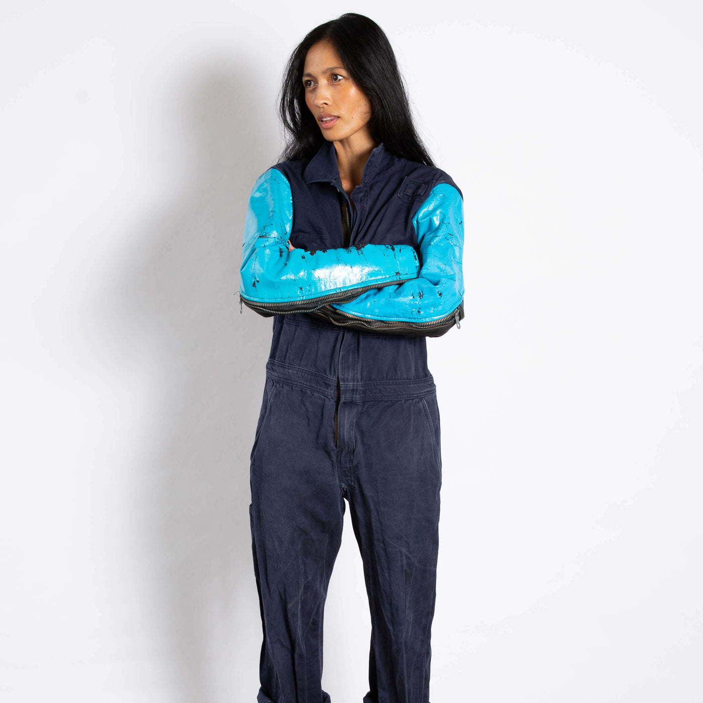 Model wears a jumpsuit that is dark navy blue and zipper up the front.  the sleeves are made of a different leather materials that is bright blue.  The paint is cracked showing black showing through.  zippers on sleeves zip up to the elbows.