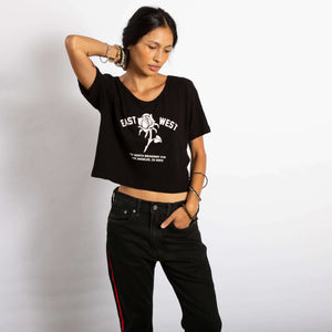 Model wears Black shirt with scooped neck and cropped length. Graphic of a rose reads "East West 727 N. Broadway #115, Los Angeles, CA 90012"