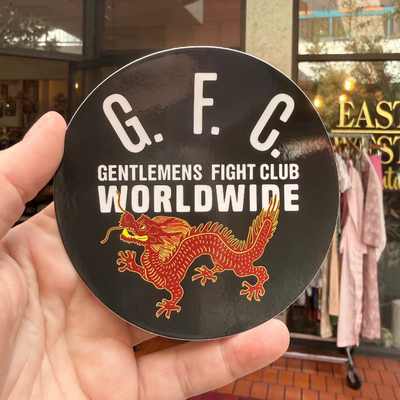 Hand holding a black sticker with 'Gentlemen's Fight Club Worldwide' and a dragon design in front of a store.