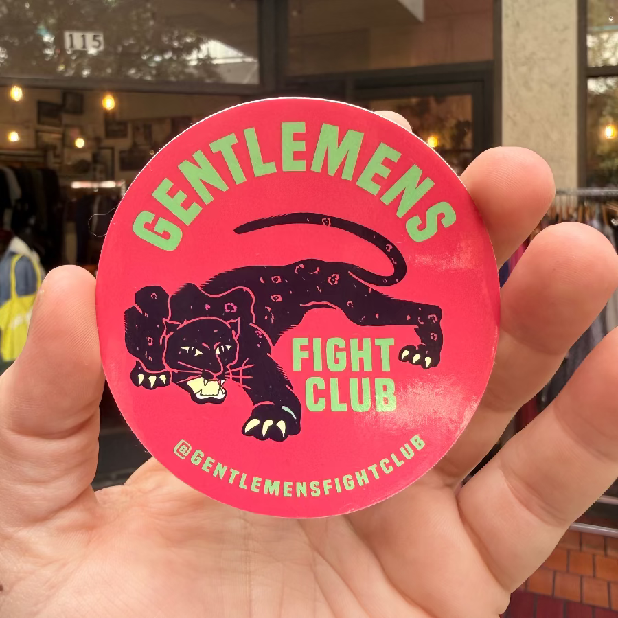 Hand holding a pink 'Gentlemen's Fight Club' sticker with a jaguar design in front of a store window.