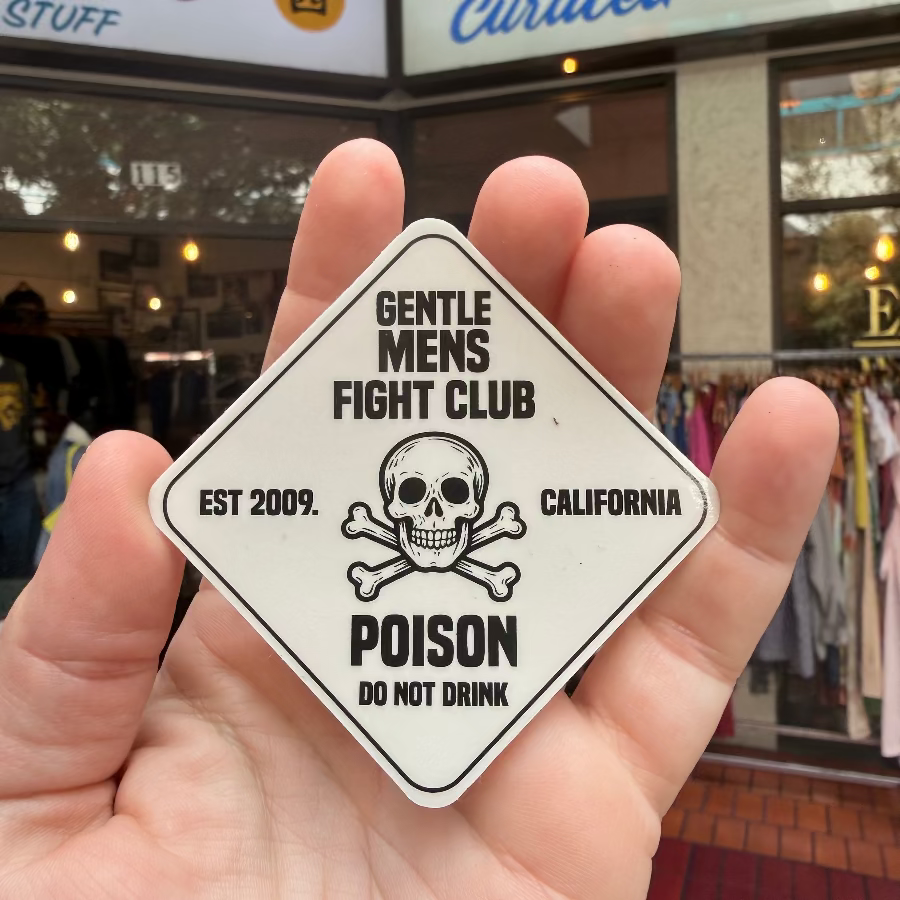 Hand holding a 'Gentle Mens Fight Club' sticker with a skull design in front of a store.