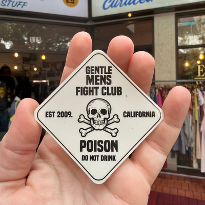 Hand holding a 'Gentle Mens Fight Club' sticker with a skull design in front of a store.