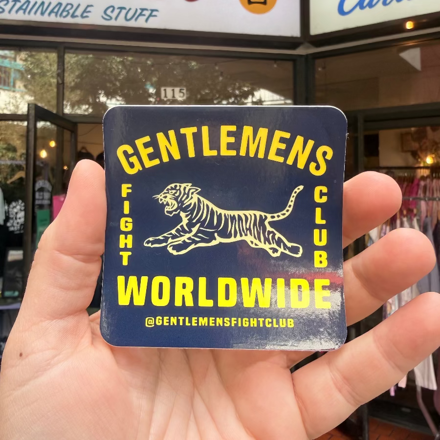 Hand holding a 'Gentlemen's Fight Club Worldwide' sticker in front of a store.