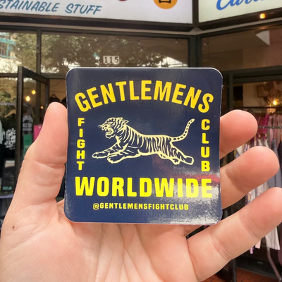 Hand holding a 'Gentlemen's Fight Club Worldwide' sticker in front of a store.