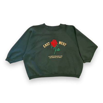 Forest green sweatshirt on white background. The sweatshirt has a red rose embroidered on the center. To the left of the rose it says “EAST” and to the right it says “WEST” under the rose reads “ 727 NORTH BROADWAY #115 LOS ANGELES, CA 90012” in pink text.