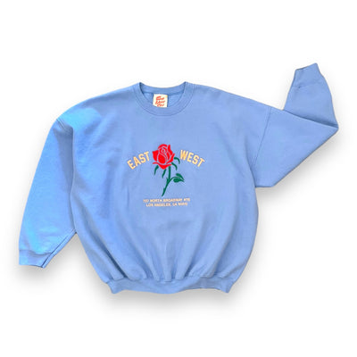 Sky blue sweatshirt on white background. The sweatshirt has a red rose embroidered on the center. To the left of the rose it says “EAST” and to the right it says “WEST” under the rose reads “ 727 NORTH BROADWAY #115 LOS ANGELES, CA 90012” in pink text.