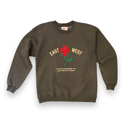 Dark speckled Heather grey sweatshirt on white background. The sweatshirt has a red rose embroidered on the center. To the left of the rose it says “EAST” and to the right it says “WEST” under the rose reads “ 727 NORTH BROADWAY #115 LOS ANGELES, CA 90012” all in pink text.