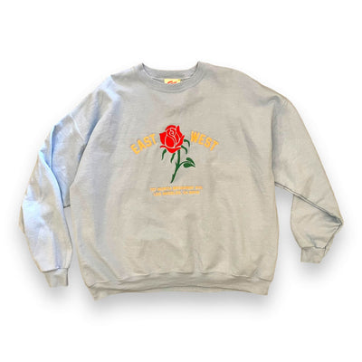 Powder blue sweatshirt on white background. The sweatshirt has a red rose embroidered on the center. To the left of the rose it says “EAST” and to the right it says “WEST” under either the rose reads “ 727 NORTH BROADWAY #115 LOS ANGELES, CA 90012”.