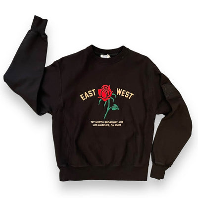 Black sweatshirt on white background. The sweatshirt has textured panels under the arms and a red rose embroidered on the center. To the left of the rose it says “EAST” and to the right it says “WEST” under the rose reads “ 727 NORTH BROADWAY #115 LOS ANGELES, CA 90012” all in pink text.