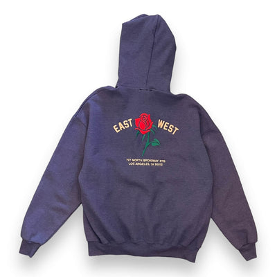 The same purpley grey zip-up hoodie from the back view on a white background. The hoodie has a red rose embroidered on the center. To the left of the rose it says “EAST” and to the right it says “WEST” under either the rose reads “ 727 NORTH BROADWAY #115 LOS ANGELES, CA 90012”.