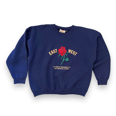 Navy sweatshirt on a white background. The sweatshirt has a red rose embroidered on the center with pink text “EAST WEST” on either side of the rose. Below the rose reads “ 727 North Broadway #115 Los Angeles CA 90012”.