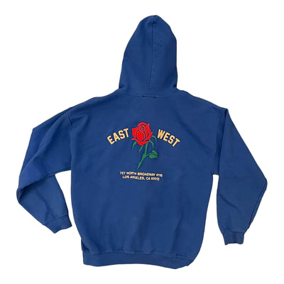 The same blue zip-up hoodie from the back view on a white background. The hoodie has a red rose embroidered on the center. To the left of the rose it says “EAST” and to the right it says “WEST” under either the rose reads “ 727 NORTH BROADWAY #115 LOS ANGELES, CA 90012”.