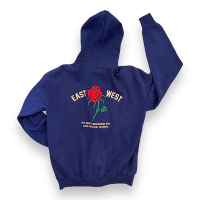 A Navy zip-up hoodie from the back view on a white background. The hoodie has a red rose embroidered on the center. To the left of the rose it says “EAST” and to the right it says “WEST” under the rose reads “ 727 NORTH BROADWAY #115 LOS ANGELES, CA 90012” in light pink text.