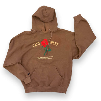 Cinnamon colored hoodie on white background. The sweatshirt has a red rose embroidered on the center. To the left of the rose it says “EAST” and to the right it says “WEST” under either the rose reads “ 727 NORTH BROADWAY #115 LOS ANGELES, CA 90012”.