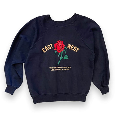 Dark Navy sweatshirt on white background. The sweatshirt has a red rose embroidered on the center. To the left of the rose it says “EAST” and to the right it says “WEST” under the rose reads “ 727 NORTH BROADWAY #115 LOS ANGELES, CA 90012” all in pink text.